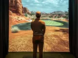 Man playing on an indoor golf simulator, standing on a mat and facing a realistic desert-themed virtual course displayed on a large screen.
