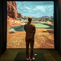 Man playing on an indoor golf simulator, standing on a mat and facing a realistic desert-themed virtual course displayed on a large screen.