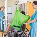 Two healthcare workers use a green patient lift sling to safely transfer a patient from a hospital bed to a wheelchair in a medical facility.