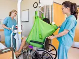 Two healthcare workers use a green patient lift sling to safely transfer a patient from a hospital bed to a wheelchair in a medical facility.
