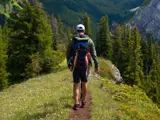 Man hiking on a narrow mountain trail surrounded by pine trees, wearing a backpack and outdoor gear, with scenic forested valleys and cliffs in the background.