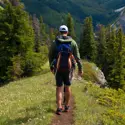 Man hiking on a narrow mountain trail surrounded by pine trees, wearing a backpack and outdoor gear, with scenic forested valleys and cliffs in the background.