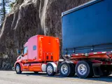 Orange semi-truck with a black trailer drives along a highway next to a rocky cliffside, highlighting industrial transport and heavy-duty logistics in action.