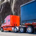 Orange semi-truck with a black trailer drives along a highway next to a rocky cliffside, highlighting industrial transport and heavy-duty logistics in action.