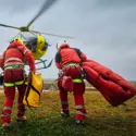 Two rescue workers in red safety gear carry equipment toward a yellow helicopter on a grassy field, preparing for an emergency medical evacuation mission.