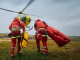 Two rescue workers in red safety gear carry equipment toward a yellow helicopter on a grassy field, preparing for an emergency medical evacuation mission.